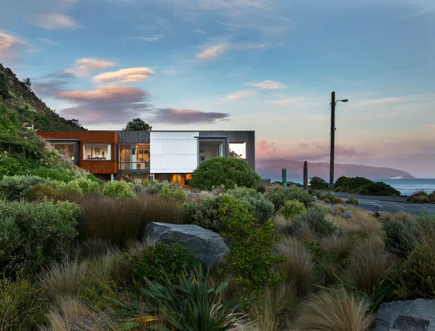 Exterior view of a contemporary multi-level home set into a lush coastal landscape. The building has crisp white and dark cladding, extensive glazing, and warm timber-toned sections, with a rocky vegetated hillside on one side and ocean views beyond. Native grasses, shrubs, boulders, and a simple driveway or access road surround the house under a dramatic blue-and-pink sky.