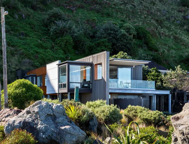 Exterior view of a contemporary multi-level hillside dwelling set among rugged rocks and dense native vegetation. The home features dark vertical cladding, warm timber-toned panels, extensive glazing, projecting box-like volumes, a flat roof, and a glass-balustraded upper terrace. An older corrugated-roof structure is visible beside it.