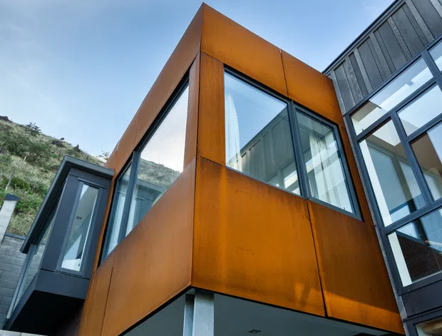 Low-angle exterior view of a contemporary multi-level building with projecting box-like volumes, large glazed openings, dark metal framing, and a prominent rust-toned weathered steel facade. The structure is set against a steep vegetated hillside.