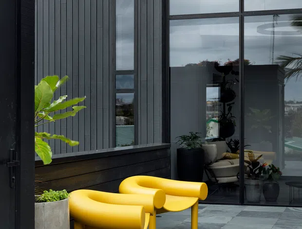A contemporary dark-clad building entrance or terrace with tall black-framed glazing, vertical charcoal cladding, grey stone paving, and two sculptural bright yellow chairs. Large planters and indoor plants soften the otherwise monochromatic facade, while the glazing reflects and reveals an interior seating area.