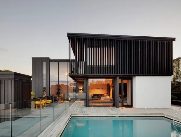 Exterior view of a contemporary multi-level residence with a white rendered lower level and a dark upper volume screened by closely spaced vertical battens. Glazed doors open toward a timber deck and swimming pool, with glass balustrades and warm interior lighting visible inside. The composition highlights the building facade, pool terrace, and strong horizontal and vertical architectural lines.