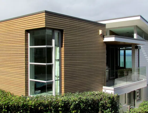 Contemporary multi-level coastal house viewed from outside, with a boxy dark timber-clad volume paired with a light horizontal-clad wing. Floor-to-ceiling glazing, projecting balconies, and a broad low-pitched roof are visible, with ocean water seen beyond the building.