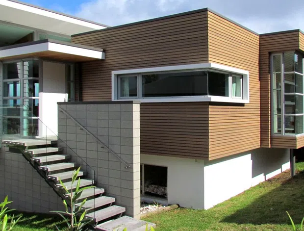 Exterior view of a contemporary multi-level house with projecting box-like forms, horizontal timber-look wall cladding, white rendered sections, extensive glazing, and an open external stair beside a grey masonry privacy wall. The building sits on a sloped grassy site under a partly cloudy sky.