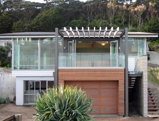 Front exterior view of a contemporary multi-level house set against dense vegetation. The facade combines broad glazed sections, pale rendered or masonry walls, dark structural framing, a timber-clad garage door and upper balcony, with a prominent pergola and exposed external stairway.