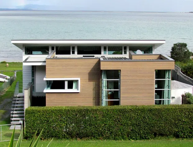 A modern coastal waterfront residence viewed from the front, with horizontal timber cladding, broad flat roof planes, extensive clerestory-style glazing, and a manicured hedge in the foreground. The house sits directly beside calm water, with a narrow lawn, steps, and a small boat visible on the site.
