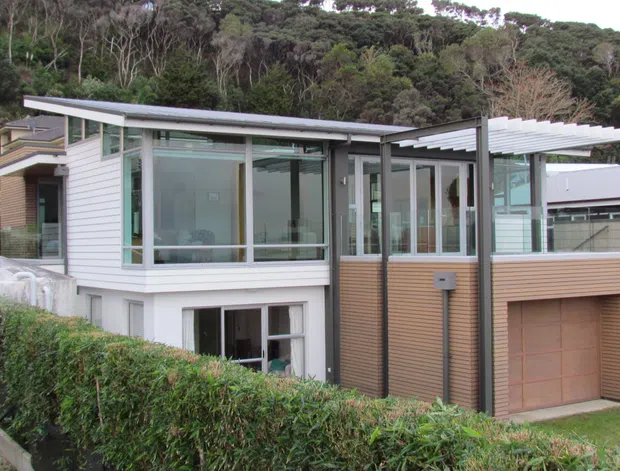Exterior view of a contemporary multi-level residence set against a wooded hillside. The home combines light horizontal weatherboard cladding, extensive glazing, a low-pitched roof, a projecting upper-level living area, and a timber-toned garage volume with a pergola-like roof structure. A manicured hedge and sloping site frame the foreground.