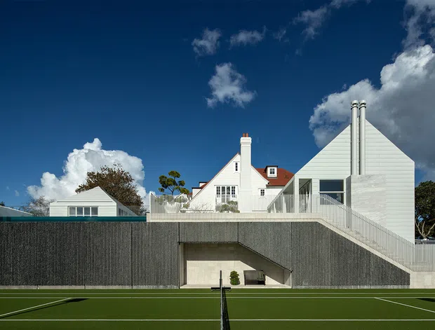 Exterior architectural view of a contemporary white-clad residential property seen beyond a tall textured boundary wall and tennis court. The composition includes a steep gabled main volume, a white weatherboard-like facade, tall chimney elements, extensive glazing, and a prominent elevated terrace or stair structure. The image is a polished architectural photograph under a vivid blue sky.