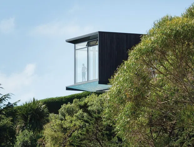 A partially concealed modern building volume rises above dense mature landscaping. The visible upper structure has dark vertical cladding, a flat roof, and a projecting corner of floor-to-ceiling glazing with a slim dark frame. Blue sky and trees surround the building, suggesting a green suburban or landscaped site.