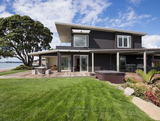 Two-storey contemporary coastal home with dark horizontal cladding, a shallow sloping roof, extensive glazing, and a large covered outdoor terrace. The house sits beside a landscaped lawn with timber decking, a spa, planting, and a visible beach or waterfront beyond.