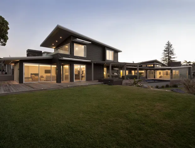 Wide exterior view of a contemporary multi-level residence at dusk, with dark horizontal cladding, broad roof overhangs, extensive warm-lit glazing, timber decking, and a connected lower wing arranged around a large lawn.