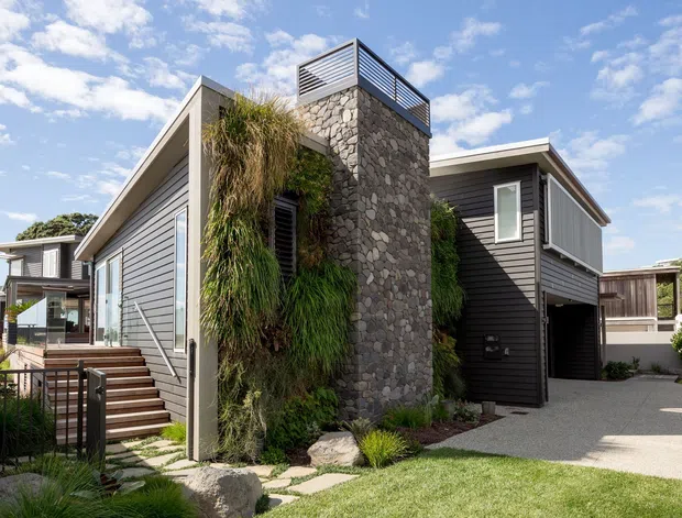 Contemporary multi-level residential exterior with dark horizontal cladding, a prominent natural-stone tower, rooftop metal screening, and integrated vertical landscaping. Timber steps lead to a covered entry and elevated deck, while a paved driveway and lawn sit in front of the house under a bright sky.