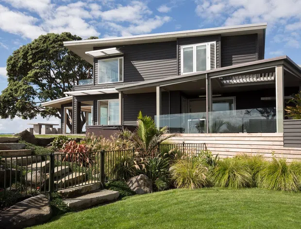 Exterior view of a contemporary two-storey home with dark horizontal cladding, a low-pitched roof, expansive windows, covered upper-level terrace, and glazed balcony railing. The house is set within a landscaped garden with ornamental grasses, palms, stone edging, and a pedestrian gate, under a bright partly cloudy sky.