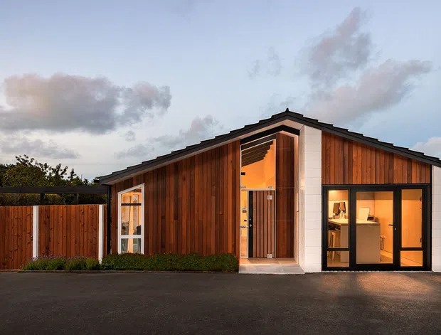 Single-storey contemporary residential building viewed from the street at dusk. The compact gabled structure features warm vertical timber cladding, crisp white rendered or panelled sections, dark metal roofing, and large glazed entry and garage doors. A matching timber boundary fence and tall narrow evergreen frame the facade.