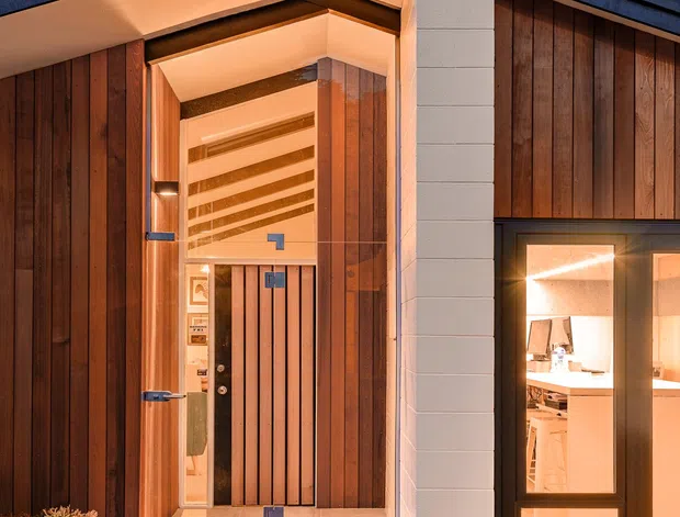 A close-up dusk view of a contemporary residential entrance. The facade combines dark vertical timber cladding, a white masonry pier, and a dark pitched roof with a projecting covered entry. Warm recessed lighting illuminates the timber-lined porch, vertical slatted front door, tiled threshold, and adjacent full-height glazed window revealing part of the interior.