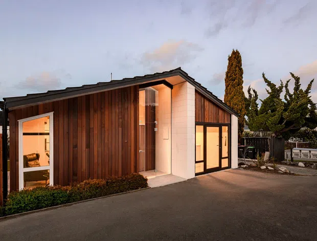 Single-storey contemporary building viewed from outside at dusk, with a sharply pitched gable roof, dark vertical timber cladding, pale fibre-cement or rendered wall panels, and large glazed openings with warm interior lighting. A paved forecourt and landscaped planting surround the compact structure.