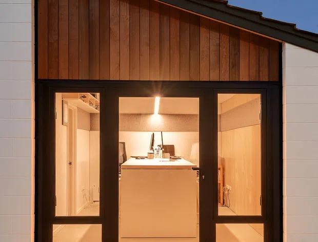 A dusk exterior view of a compact modern building or extension with a steeply sloping dark roof, white rendered or panelled side walls, and a warm vertical timber-clad gable facade. Large black-framed glazed doors reveal a brightly lit interior containing a central island or workbench and pale timber finishes.