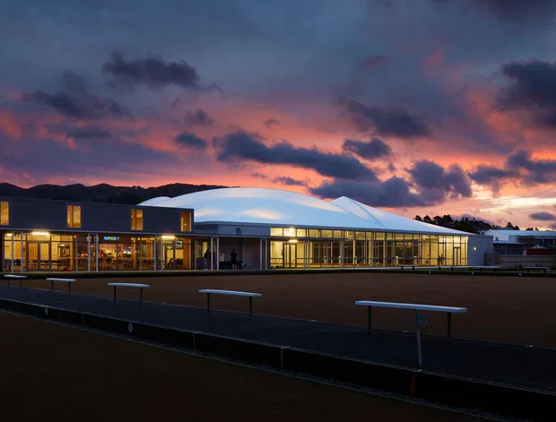 A low, contemporary institutional or commercial building photographed at dusk, with extensive illuminated glazing, dark rectangular wall volumes, and a prominent white curved and faceted roof canopy. The building fronts a broad paved or asphalt forecourt with simple bench-like rails, while a dramatic sunset sky forms the background.