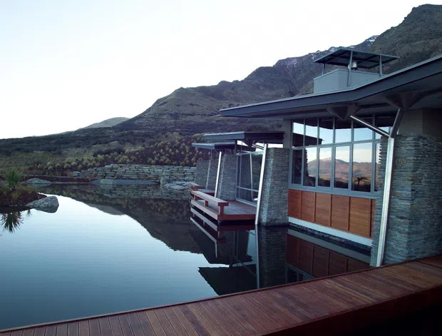 Modern multi-level lakeside residence built into a mountainous setting, with dark metal roof forms, extensive glazing, stacked-stone walls, timber-clad panels, and a projecting timber deck beside still water.