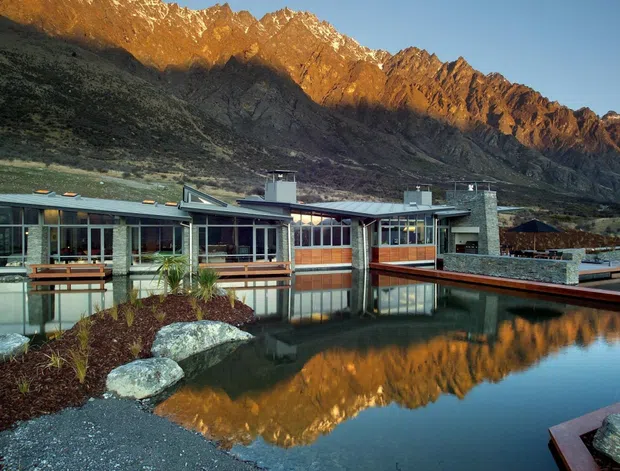 A low-slung modernist mountain residence or lodge set beside a reflecting pond, with extensive floor-to-ceiling glazing, stone walls, dark metal roof planes, and timber-framed terraces. Rugged alpine mountains rise directly behind the building and are reflected in the still water. The composition emphasises the relationship between architecture, landscape, and water, with planted rock gardens and narrow projecting decks in the foreground.