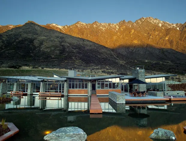 A low, contemporary mountain retreat set beside a reflective pond, with extensive glazing, flat roof forms, timber decking, and stone-clad elements. Rugged alpine mountains dominate the background, catching warm late-day light and reflecting in the water.