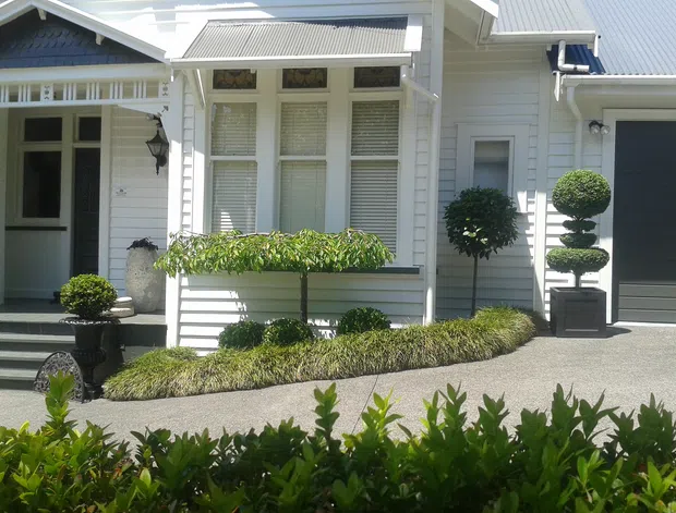 Front exterior of a refined white weatherboard home with a gabled roof, decorative window detailing, covered entry, garage, and formal clipped topiary landscaping along a paved driveway. The facade combines traditional residential character with carefully maintained contemporary landscaping.