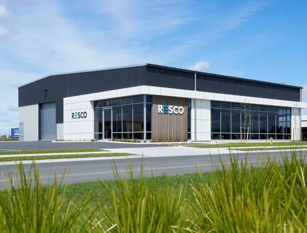 A modern single-storey commercial building photographed from the roadside, with a long rectangular form, dark corrugated upper facade and roof, white panel cladding, expansive glazed frontage, and a timber-look entrance feature bearing the RESCO logo. Landscaped grasses and a paved access road occupy the foreground.
