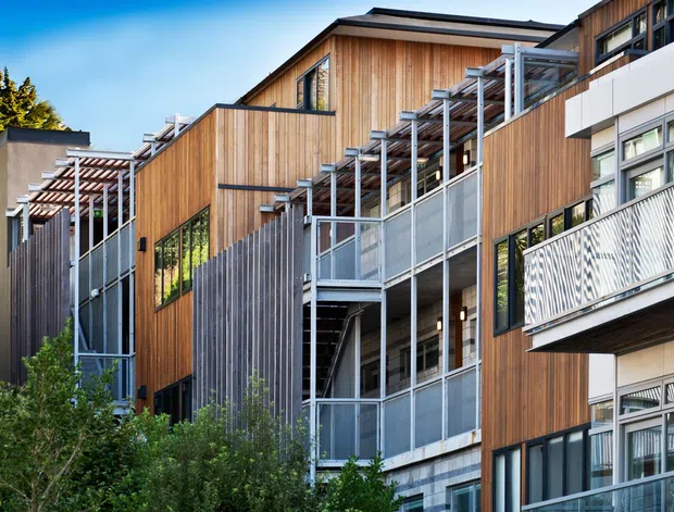 Exterior view of a contemporary multi-storey residential building with extensive warm timber cladding, dark-framed glazing, projecting balconies, metal balustrades, and repeated pergola-like structural screens. The stepped facade is integrated into a leafy urban or suburban setting.