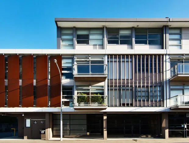 Exterior view of a contemporary multi-storey building with a flat roof, broad horizontal glazing, projecting balconies, vertical metal screening, and warm reddish-brown facade panels. The ground level includes recessed glazed entries and dark service or parking openings.