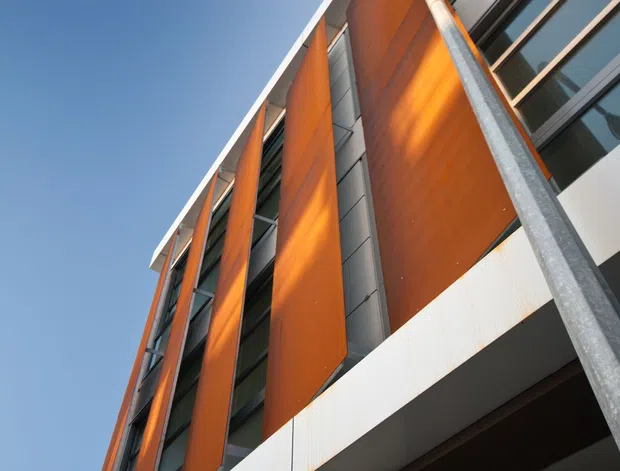 Upward-looking exterior view of a contemporary multi-storey building facade, featuring tall orange-brown vertical cladding panels, dark metal-framed glazing, white horizontal soffit or balcony bands, and prominent galvanized steel structural posts against a clear blue sky.