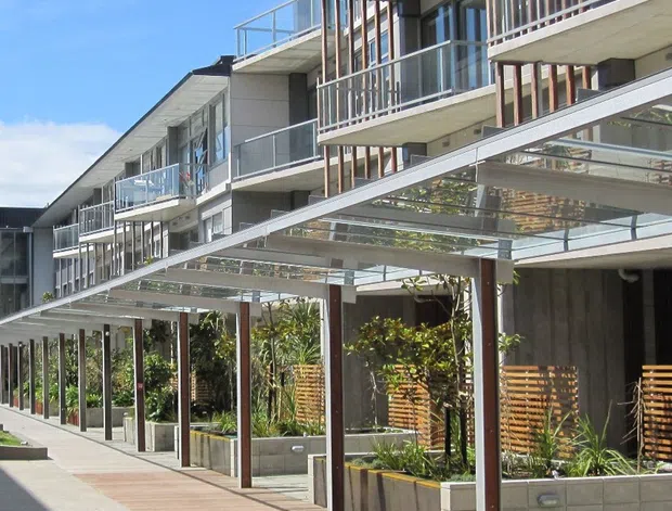 Exterior view of a contemporary multi-storey apartment building with repeated projecting balconies, clear glass balustrades, slender timber-toned vertical screens, and a covered pedestrian walkway/pergola at ground level. The frontage combines light rendered panels, exposed concrete balcony slabs, dark framing, timber decking, raised concrete planters, and landscaped planting.