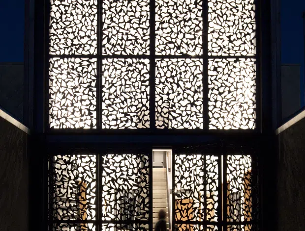Night-time view of a narrow, multi-storey contemporary building facade. Large illuminated patterned screens with irregular perforations cover the upper glazing and flank the ground-floor entrance, creating a dramatic glowing geometric effect. Dark vertical cladding, black framing, and a paved entry court emphasize the high-contrast architectural composition.