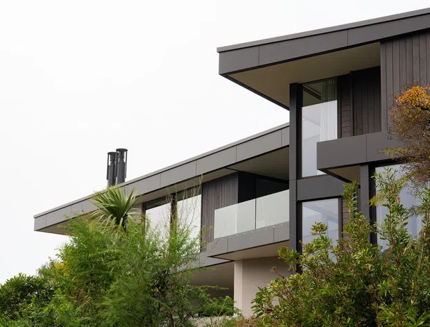 Exterior view of a contemporary multi-level residence with strong horizontal roof planes, dark vertical cladding, expansive glazing, and glass balcony balustrades. Landscaping partially screens the lower level, while a chimney or flue is visible on the roofline.