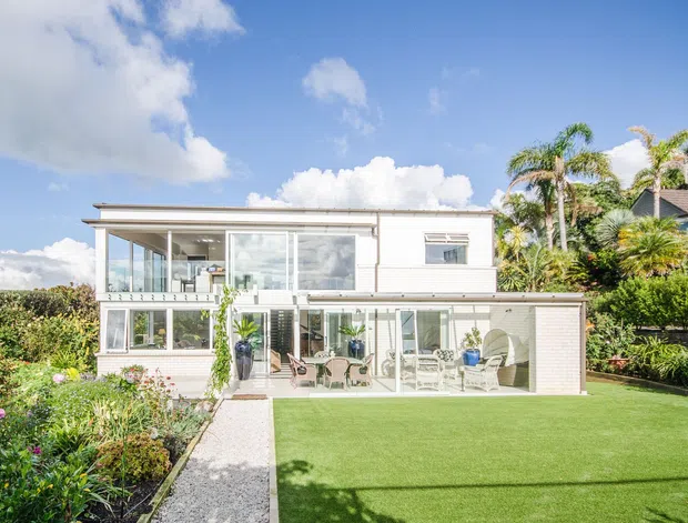 Bright exterior view of a two-storey contemporary residential house with a white rendered facade, extensive glazing, balconies and terraces, and a flat roofline. The property is surrounded by manicured hedges, palm trees, flowering garden beds, gravel paths, and a neatly maintained lawn.