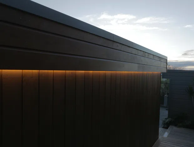 Close exterior view of a contemporary flat-roof structure with dark horizontal wall cladding above vertical timber-like screening. Warm concealed linear lighting illuminates the junction between the upper wall and lower screen, with a small timber deck and landscaped courtyard visible at the side. The composition has strong horizontal lines and a moody dusk atmosphere.