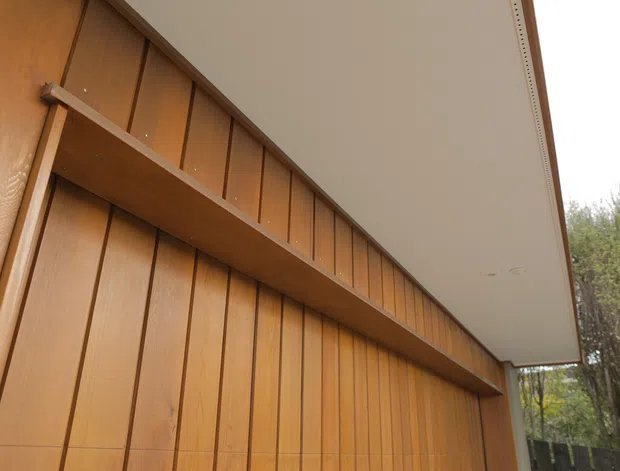 Close-up architectural view of a warm timber-clad exterior wall with narrow vertical boards and an integrated horizontal shelf or canopy detail beneath a broad smooth roof overhang. The scene appears to be outdoors, with trees visible beyond the structure.