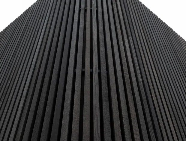 Close-up upward view of a dark, vertically ribbed architectural facade composed of closely spaced timber-like slats, curving toward a rounded roofline or apex against a plain white sky. The image emphasizes strong repetition, texture, and geometric form.