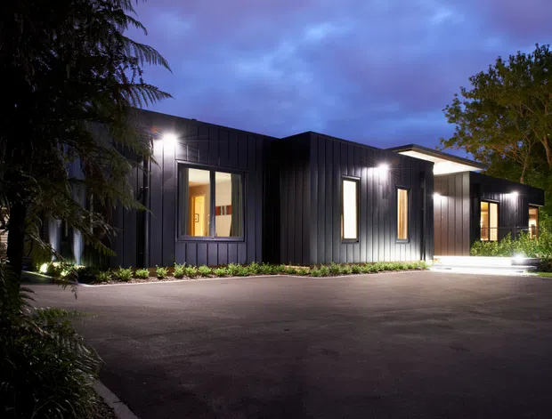 Twilight exterior view of a low-rise contemporary building with dark vertically ribbed cladding, illuminated windows and entry, and a broad paved driveway. Dense landscaped planting frames the building, while warm interior light contrasts with the cool evening sky.