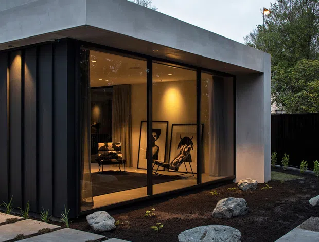 Exterior view of a compact contemporary pavilion or extension with a smooth grey concrete-like flat roof and deep overhang, dark vertical cladding, and large black-framed floor-to-ceiling glazing. Warm interior lighting reveals a lounge-like space with framed artwork and lounge chairs. The building is set within a minimalist gravel-and-rock landscape with large concrete stepping slabs and planted borders.
