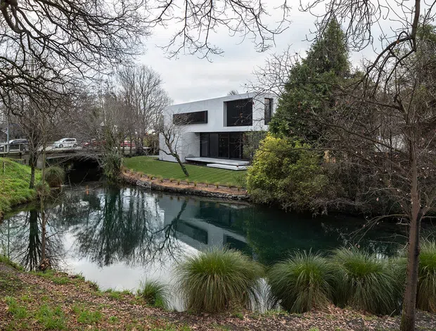 Exterior view of a contemporary two-storey residence beside a landscaped pond or stream, framed by mature deciduous trees and dense planting. The house has a minimalist white rendered facade, dark expansive window openings, and a flat roof. A small bridge and driveway are visible to the left, with the water and reflections dominating the foreground.
