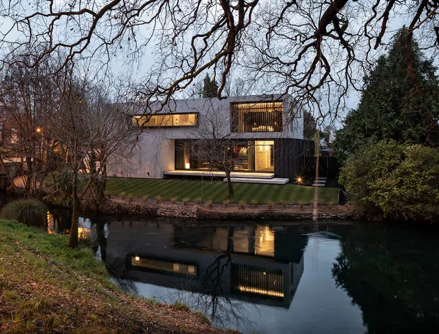 A contemporary two-storey residence viewed across a tranquil pond at dusk. The house has a rectilinear flat-roofed form, pale rendered walls, extensive dark-framed glazing, warm interior lighting, and screened upper-level outdoor spaces. Mature trees and dense planting surround the waterfront setting, with the building reflected in the water.