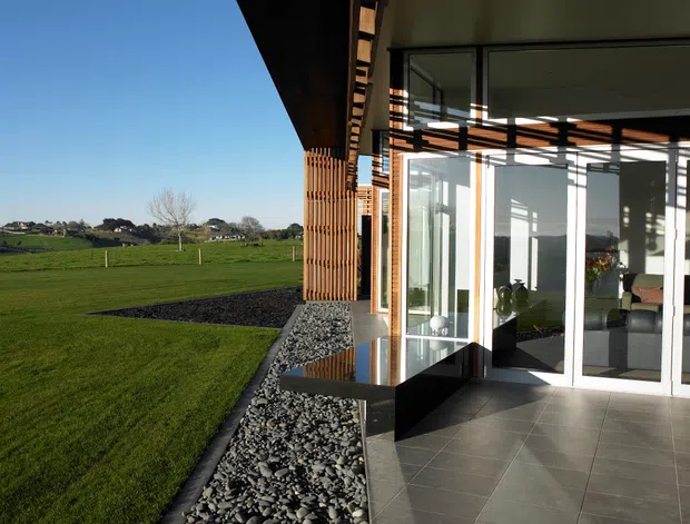 Exterior view of a contemporary pavilion-like building beside an open grassy rural landscape. The image focuses on a covered terrace or veranda with large floor-to-ceiling glazed doors and windows, dark grey tiled paving, a pebble drainage strip, timber screening, and strong linear roof and framing details. Interior furnishings are visible through the glazing.