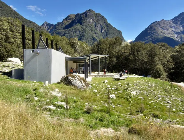A small contemporary building set in a rugged alpine mountain landscape. The structure combines a raw concrete service volume with a lightweight dark metal-framed canopy and extensive open glazing. Rocky grassland, native trees, steep forested mountains, and a clear blue sky surround the building, with a narrow driveway or path visible at right.