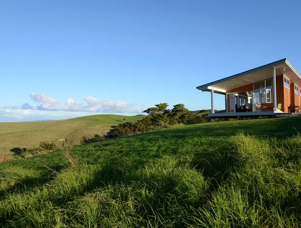A contemporary single-storey house sits on a grassy coastal or rural hillside, with broad glazed openings, a projecting flat roof, and warm timber-toned wall cladding. The surrounding rolling landscape and open sky dominate the setting.
