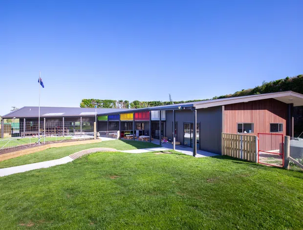 Exterior view of a low, single-storey institutional or community building arranged around a landscaped lawn and paved paths. The building has dark blue-grey cladding, a prominent timber-toned wall section, broad low-pitched rooflines, colourful window and door panels, and a fenced outdoor area. A flagpole and covered veranda-like circulation zone are visible.