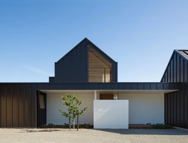 Exterior architectural photograph of a contemporary multi-volume house under a clear blue sky. The building has dark vertical metal cladding, steep gabled roof forms, a low horizontal connecting wing, and a recessed entry courtyard with a white rendered wall, small planted tree, and gravel forecourt. Timber-like horizontal screening is visible within the central gable.