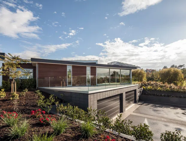 Exterior view of a contemporary, low-profile residential building set within landscaped gardens. The house features extensive floor-to-ceiling glazing, dark horizontal cladding, warm timber wall panels, a flat roof, and a raised terrace above a garage or lower-level structure. A driveway and planted garden beds occupy the foreground.