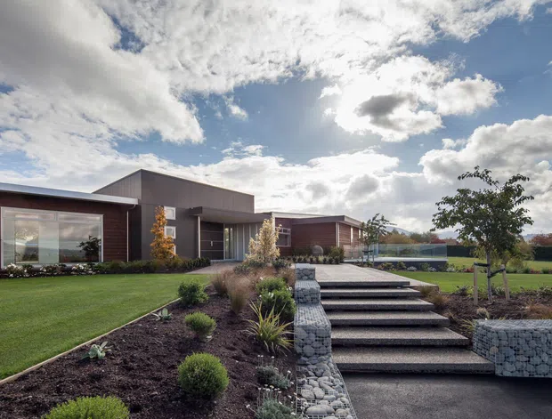 A contemporary single-storey home with dark horizontal cladding, broad glazing and low-pitched roof forms, viewed from a landscaped front garden. Broad steps lead up through planted beds and gabion retaining walls toward the entrance, with open lawn and a sunny rural-looking setting beyond.