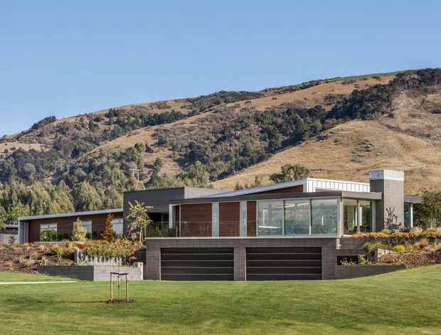 A contemporary single-storey hillside residence with a low, elongated form, dark horizontal cladding, extensive floor-to-ceiling glazing, and a prominent rectangular chimney or vertical masonry feature. The house is set into a landscaped lawn and garden, with a double garage integrated beneath the main living level. Dry, rugged hills rise directly behind the property.