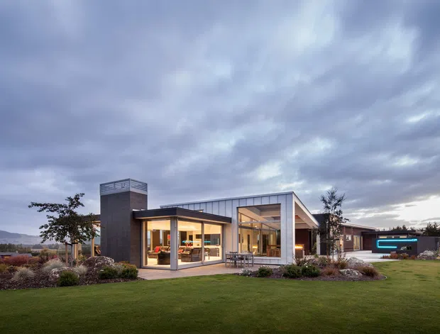 Wide exterior view of a low, modern single-storey residence set within a landscaped lawn. The home features broad floor-to-ceiling glazing, a projecting flat-roofed living wing, a taller dark-clad vertical volume with a screened top, and a pitched or sloping roof over the central glazed section. A separate dark outbuilding is visible to the right, with distant hills and a dramatic overcast sky in the background.