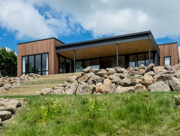 Single-storey contemporary house viewed from the landscaped front, with a low-pitched roof, extensive dark-framed glazing, warm vertical timber cladding, and a broad covered outdoor terrace. The house is set into a gently sloping site with large natural boulders, stone steps, and native-looking grass planting.
